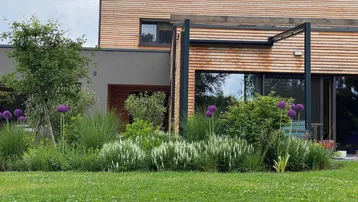 Modernes Haus mit Holzfassade, großen Fenstern und einem gepflegten Garten mit vielfältiger Bepflanzung.