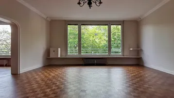 Helles Wohnzimmer mit Parkettboden, Stuckdecke und Blick ins Grüne durch große Fenster.