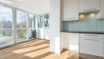 Helles Wohnzimmer mit Holzboden, großen Fenstern und integrierter Küchenzeile mit Blick ins Grüne.