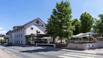 Außenansicht eines Gebäudes mit Restaurant und Metzgerei, davor eine belebte Straßenszene und eine große Außenterrasse.