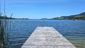 Holzsteg führt in einen klaren See unter blauem Himmel mit grünen Hügeln im Hintergrund.