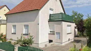 Einfamilienhaus mit rotem Ziegeldach, heller Fassade und grünem Balkon in ländlicher Umgebung.
