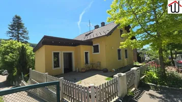 Gelbes Einfamilienhaus mit Satteldach und kleinem gepflasterten Bereich im Vorgarten.