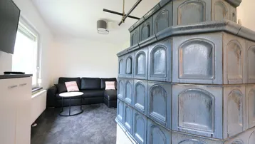 Gemütliches Wohnzimmer mit einem traditionellen Kachelofen, schwarzem Sofa und Blick ins Grüne.