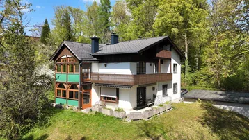 Freistehendes Haus mit Balkon und Wintergarten, umgeben von viel Grün und Bäumen.