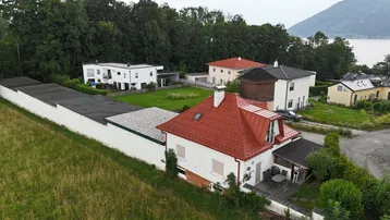 Luftaufnahme eines Wohnhauses mit rotem Dach, umgeben von Grünflächen und weiteren Gebäuden, mit See im Hintergrund.