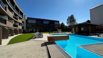 Moderner Außenbereich mit großem Pool, Liegestühlen und umliegenden Gebäuden unter strahlendem Himmel.