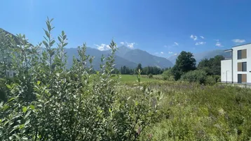 Sonniger Ausblick auf eine weite grüne Landschaft mit majestätischen Bergen und einem modernen Wohngebäude.