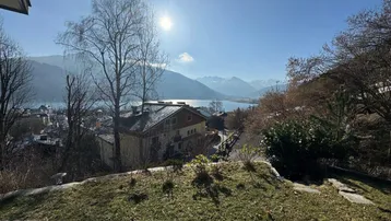 Sonniger Ausblick auf einen See und die umliegenden Berge mit einem Haus im Vordergrund und grüner Wiese.