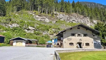 Traditionelles Steinhaus mit Parkplatz und grüner Wiese vor einer bewaldeten Bergkulisse unter blauem Himmel.