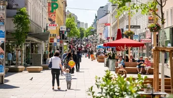 Eine belebte Fußgängerzone mit vielen Geschäften, Restaurants und Menschen, die das sonnige Wetter genießen.