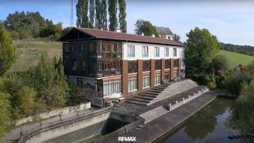 Großes Gebäude mit einzigartiger Fassade und Holzdeck am Wasser, umgeben von grüner Landschaft.