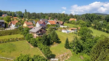 Luftaufnahme einer Wohnsiedlung mit mehreren Häusern und viel Grün in hügeliger Landschaft.