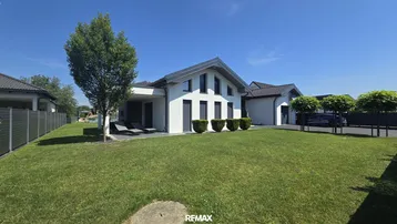 Vorderansicht des modernen Einfamilienhauses mit gepflegtem Rasen, Bäumen und einer Terrasse mit Liegestühlen.