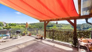 Große Terrasse mit rotem Sonnensegel, Grillplatz und weitem Panoramablick über die grüne Landschaft.