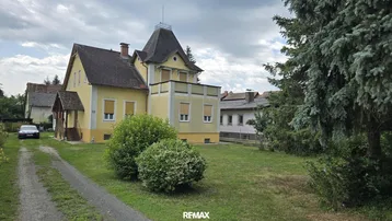 Gelbes Einfamilienhaus mit markantem Turm und großem Vorgarten, umgeben von Bäumen.