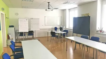 Heller Konferenzraum mit mehreren Tischen, Stühlen, Whiteboards und einem Deckenprojektor für Präsentationen.