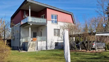 Modernes Wohnhaus mit weißer und roter Fassade, Balkon mit Glasgeländer und einem gepflegten Garten.