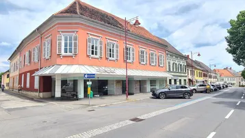 Historisches Gebäude mit rosafarbener Fassade, großen Schaufenstern im Erdgeschoss und parkenden Autos auf der Straße.