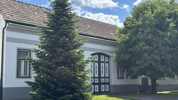 Traditionelles Haus mit weißer Fassade, grünen Fensterrahmen und einem großen Nadelbaum im Vorgarten.