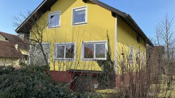 Gelbes Einfamilienhaus mit Satteldach und umgebender Vegetation an einem sonnigen Tag.