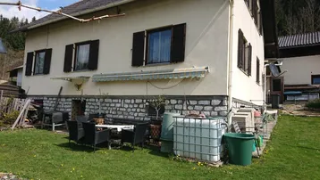 Hausfassade mit kleiner Rasenfläche, Außensitzbereich und Blick auf die umliegende Natur.