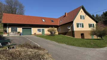 Hausfassade mit Zufahrt und Garage in ländlicher Umgebung unter blauem Himmel.