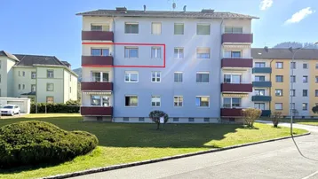 Mehrstöckiges Wohnhaus mit Balkonen, Rollläden und gepflegter Grünfläche, umgeben von Bergen.