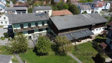 Luftaufnahme eines mehrstöckigen Gebäudes mit Balkonen und einem angrenzenden Scheunengebäude, umgeben von viel Grün.
