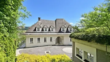 Elegante Villa mit gepflegtem Vorgarten und weitläufiger Auffahrt unter blauem Himmel.