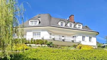 Herrschaftliche Villa mit geschwungenen Balkonen und üppiger grüner Bepflanzung in sonniger Lage.