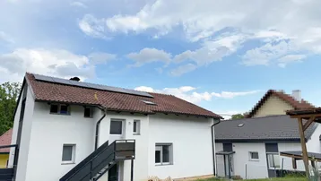 Weißes Haus mit Ziegeldach und Solaranlage, umgeben von Grünflächen unter blauem Himmel.