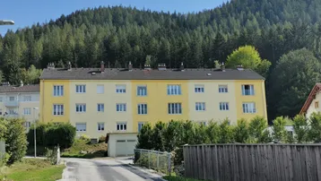 Gelbes Mehrfamilienhaus mit mehreren Etagen und einem bewaldeten Hügel im Hintergrund.