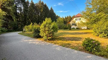 Haus in ländlicher Umgebung mit großem Garten und Wald im Hintergrund unter blauem Himmel.