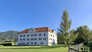Historisches Gebäude mit rotem Ziegeldach, weißer Fassade und weitläufiger Grünfläche vor Bergkulisse.
