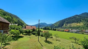 Weitläufiger Blick über ein grünes Tal mit Häusern, Bäumen und Bergen unter strahlend blauem Himmel.