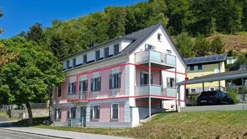 Rosa Gebäude mit mehreren Balkonen und Blick auf bewaldeten Hügel unter blauem Himmel.