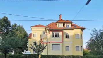 Gelbe Fassade eines Mehrfamilienhauses mit roten Dachziegeln und grüner Hecke im Vordergrund.