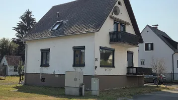 Weißes Einfamilienhaus mit Satteldach, Balkon und dunklen Fensterläden in ländlicher Umgebung.