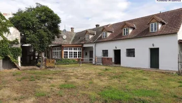 Ländliches Anwesen mit weißer Fassade, Dachgauben und großem Gartenbereich unter bewölktem Himmel.