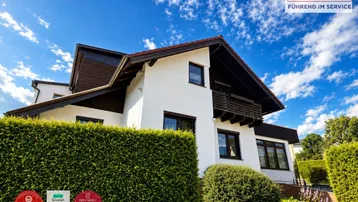 Weißes Einfamilienhaus mit Satteldach, umgeben von einer gepflegten Hecke unter blauem Himmel.