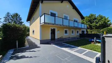 Gelbes Einfamilienhaus mit Balkon, gepflasterter Auffahrt und gepflegtem Vorgarten unter blauem Himmel.