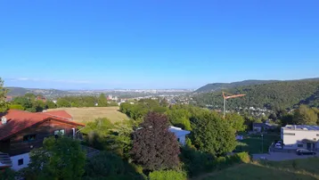 Weitläufiger Blick über eine grüne Hügellandschaft mit Stadtpanorama in der Ferne unter blauem Himmel.