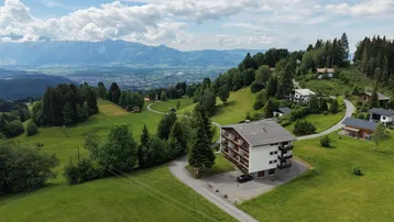Luftaufnahme des Mehrfamilienhauses in alpiner Landschaft mit weitem Blick auf das Tal und die Berge.