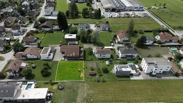 Luftaufnahme eines Wohngebiets mit einem markierten grünen Grundstück, umgeben von Häusern und Feldern.