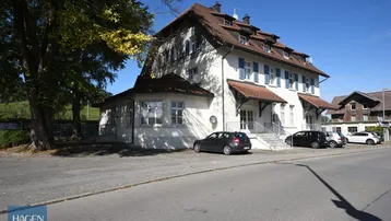 Historisches Gebäude mit weißer Fassade, blauen Fensterläden und rotem Ziegeldach an einer Straße.