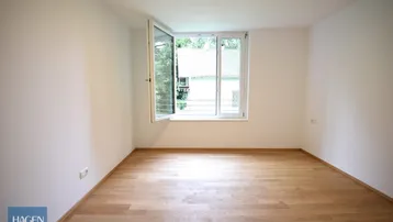 Heller, leerer Raum mit Holzboden und einem geöffneten Fenster, das Tageslicht hereinlässt.