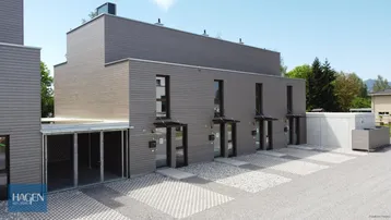 Moderne Reihenhäuser mit Holzfassade und gepflasterten Parkflächen unter blauem Himmel.