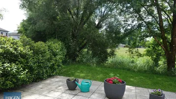 Gepflasterte Terrasse mit Blumentöpfen und Blick ins Grüne, umgeben von Bäumen und Hecken.
