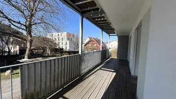 Langer, sonniger Balkon mit Holzboden und Blick auf die umliegenden Gebäude und Bäume.